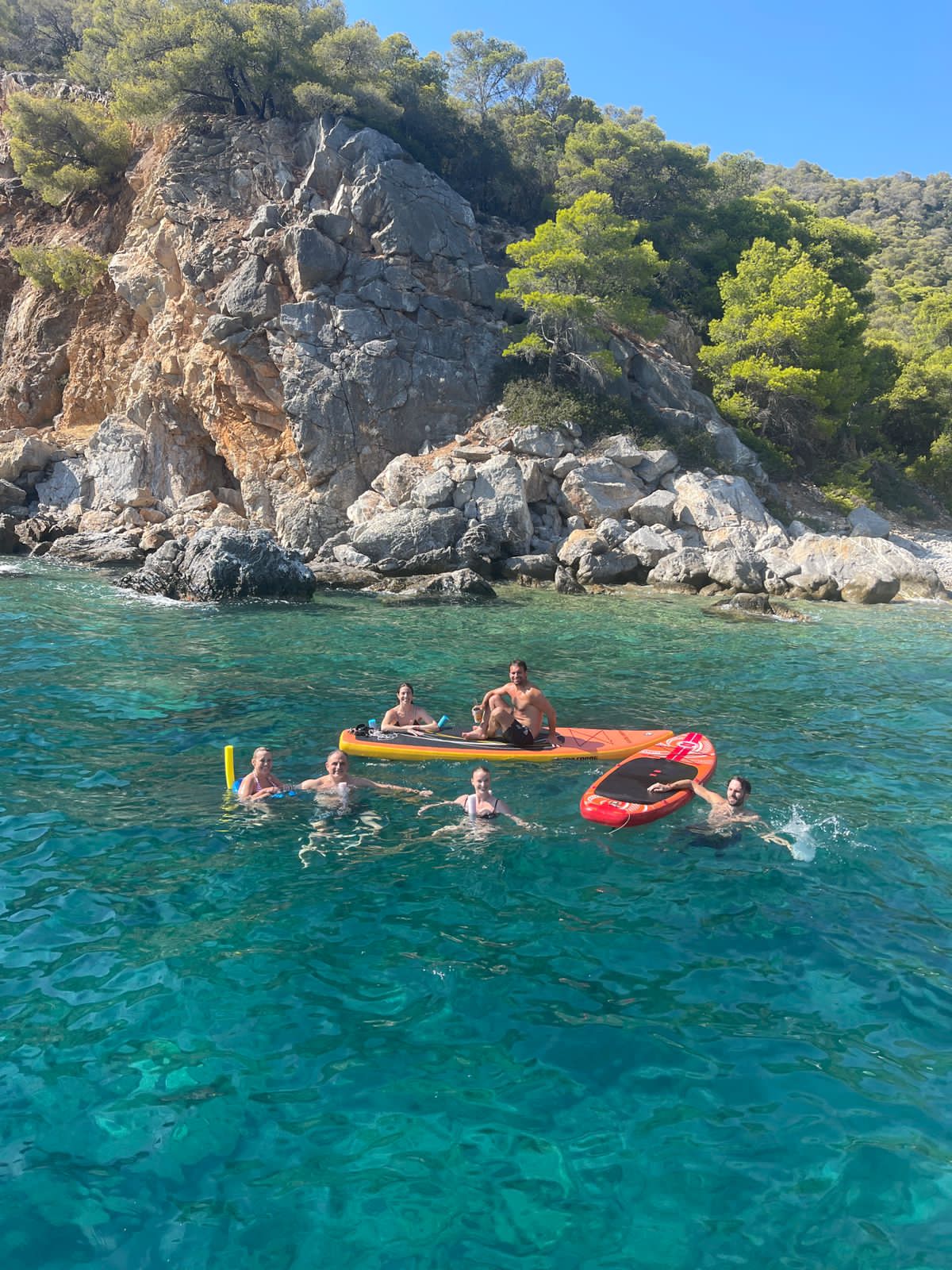 Mehrere Menschen baden während eines Segelurlaubs vergnügt im Mittelmeer in türkisem Wasser vor einem Felsen. Sie haben ein Kayaks, ein SUP und eine Schwimmnudel dabei.