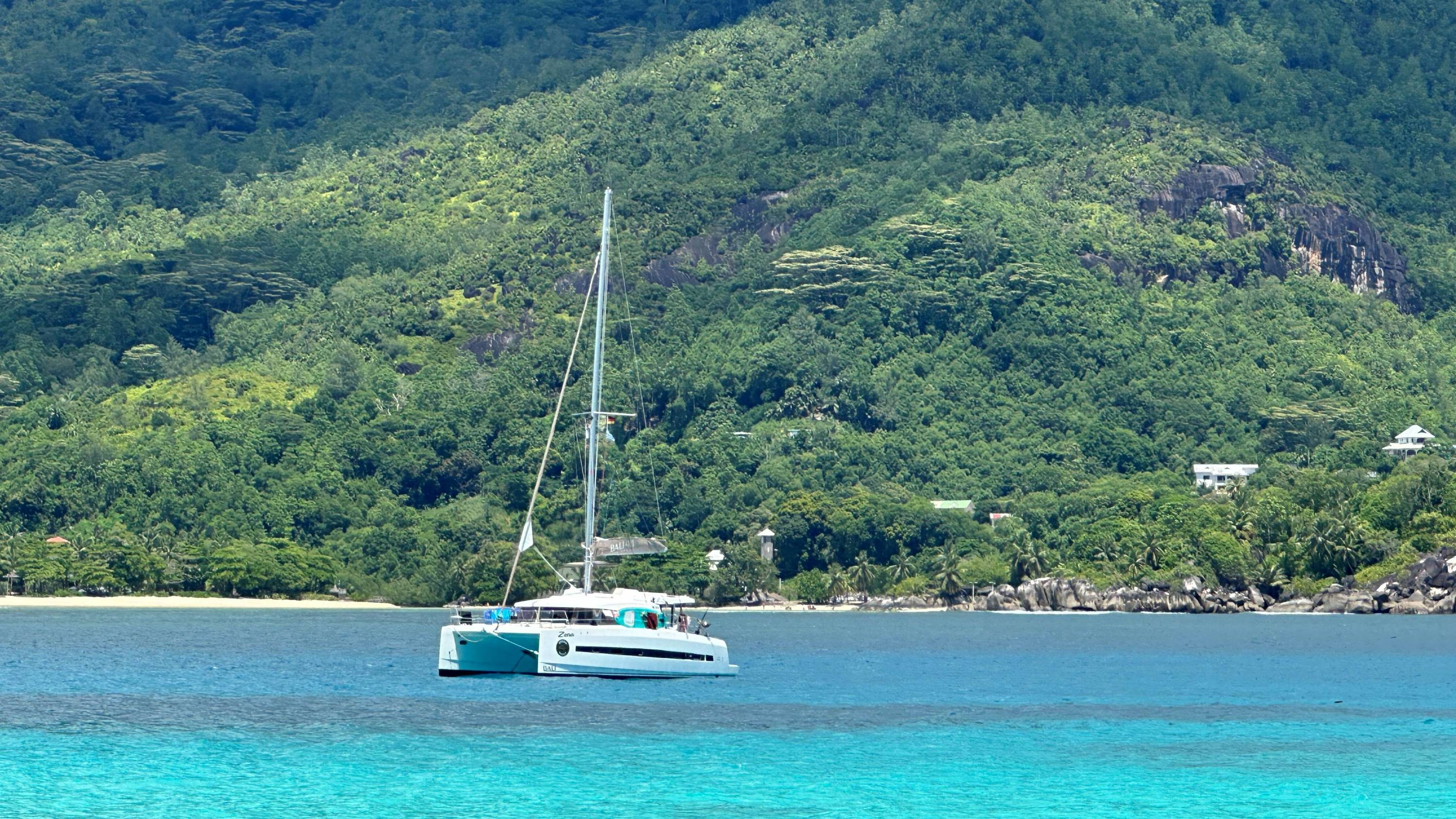Katamaran vor Anker liegend in türkisblauem Wasser vor einer grünbewachsenen Insel auf den Seychellen